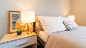 Uma cama bem arrumada com lençóis brancos, uma luminária de cabeceira, um quadro emoldurado e uma pequena planta em vaso sobre uma mesa de cabeceira.