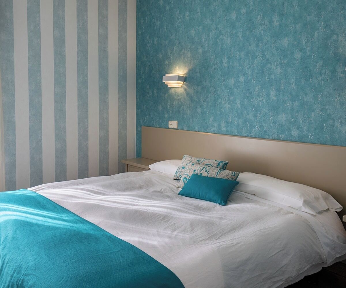 Ein Bett mit weißer Bettwäsche und einer türkisblauen Decke, ein Nachttisch und eine Wandleuchte.