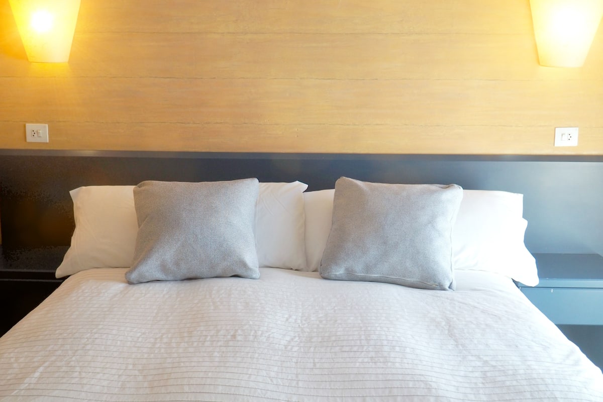 Cama bem arrumada com lençóis brancos e duas almofadas cinzas, uma cabeceira escura e duas luzes fixadas na parede.