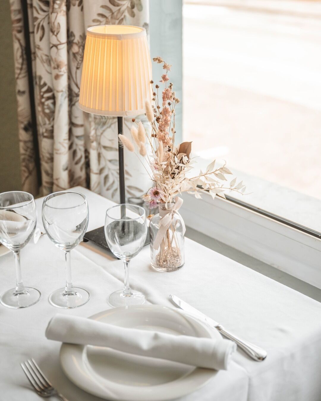 Una mesa dispuesta con tres copas de vino, un florero con flores secas, una lámpara y cubiertos.