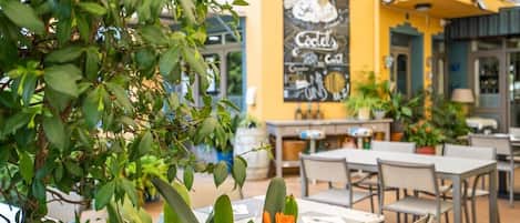 An outdoor café with tables and chairs, a yellow wall with a chalkboard menu, and potted plants.