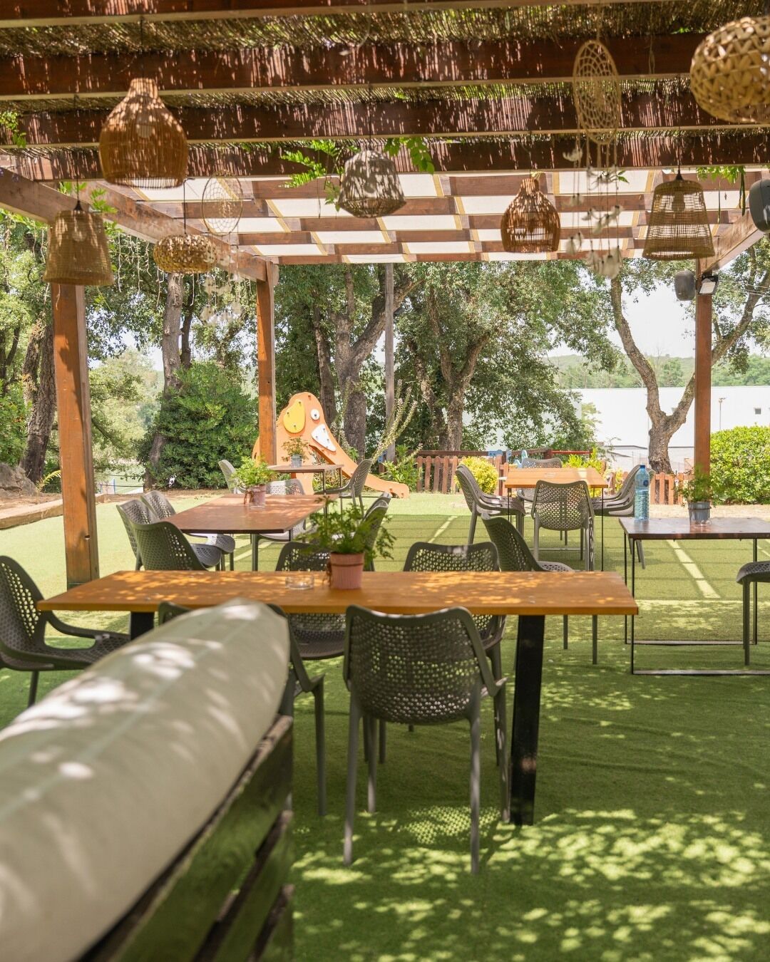 Un área de estar al aire libre con mesas y sillas, una pérgola con luces colgantes, y vistas a árboles y una masa de agua.