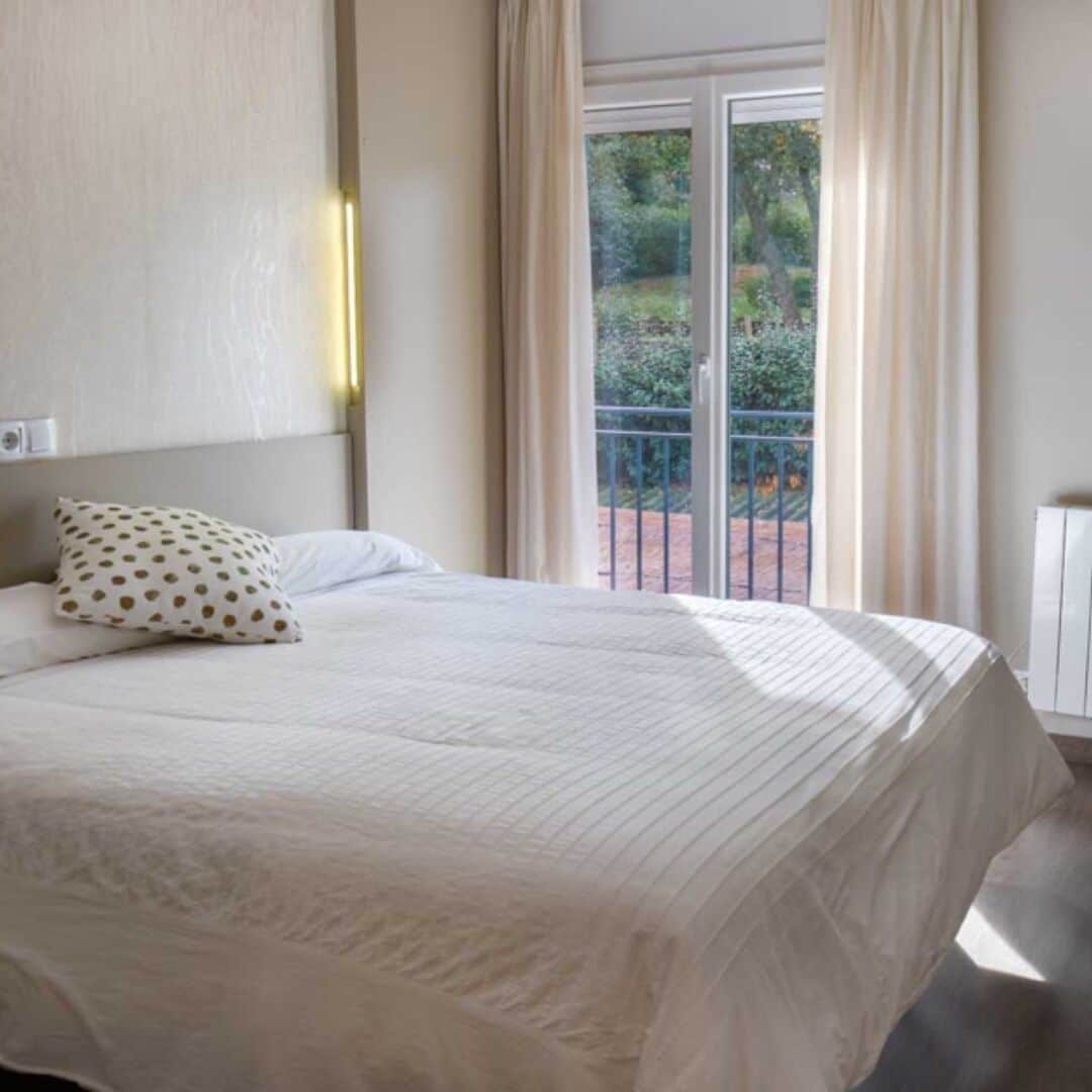 A hotel room with a bed, a pillow, a nightstand, and a view of greenery outside.