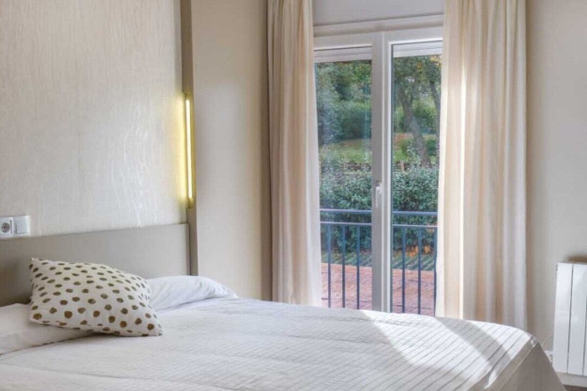 A hotel room with a bed, a pillow, a nightstand, and a view of greenery outside.