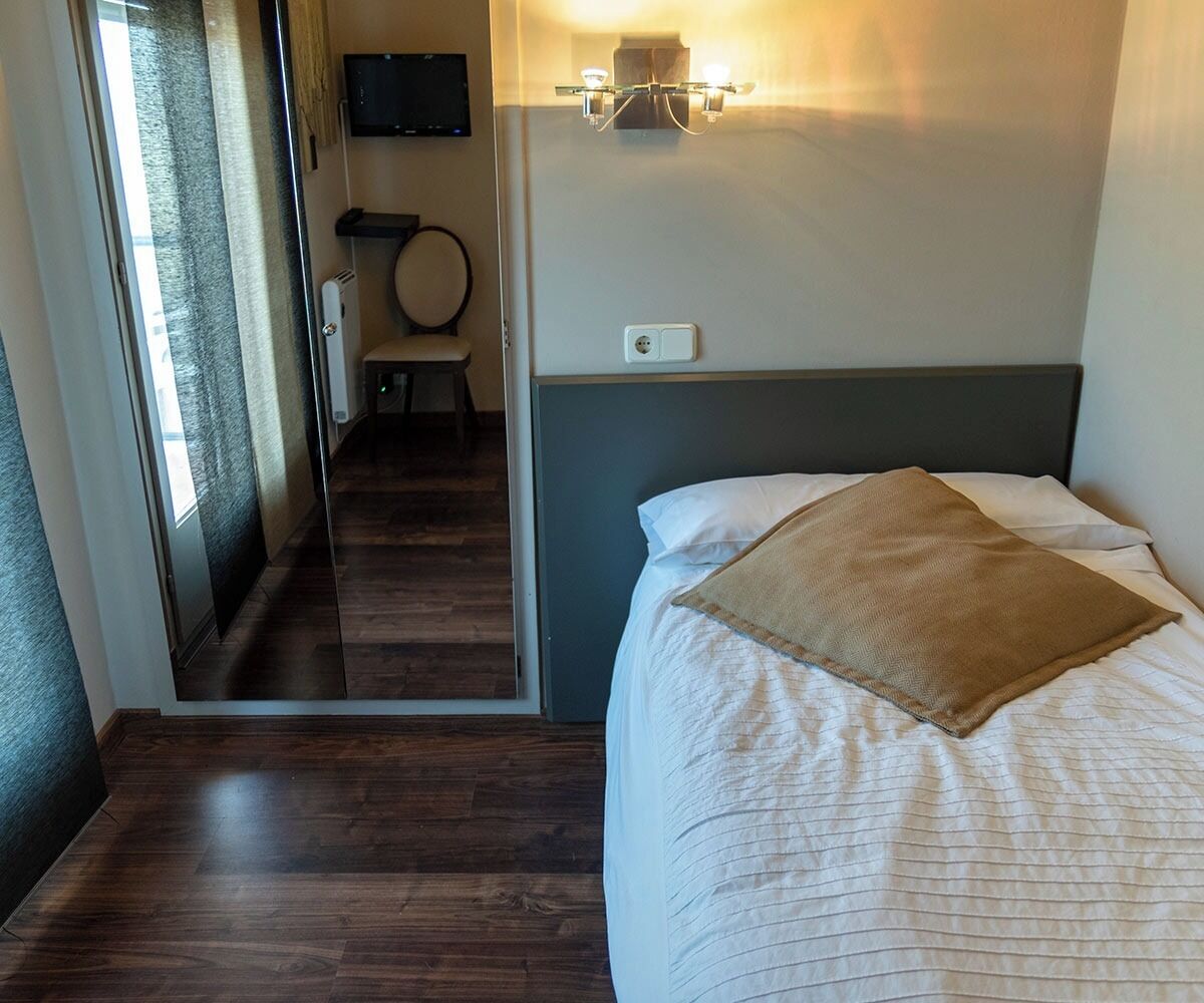 Quarto de hotel com cama, mesa de cabeceira, cadeira, televisão e luminária na parede.