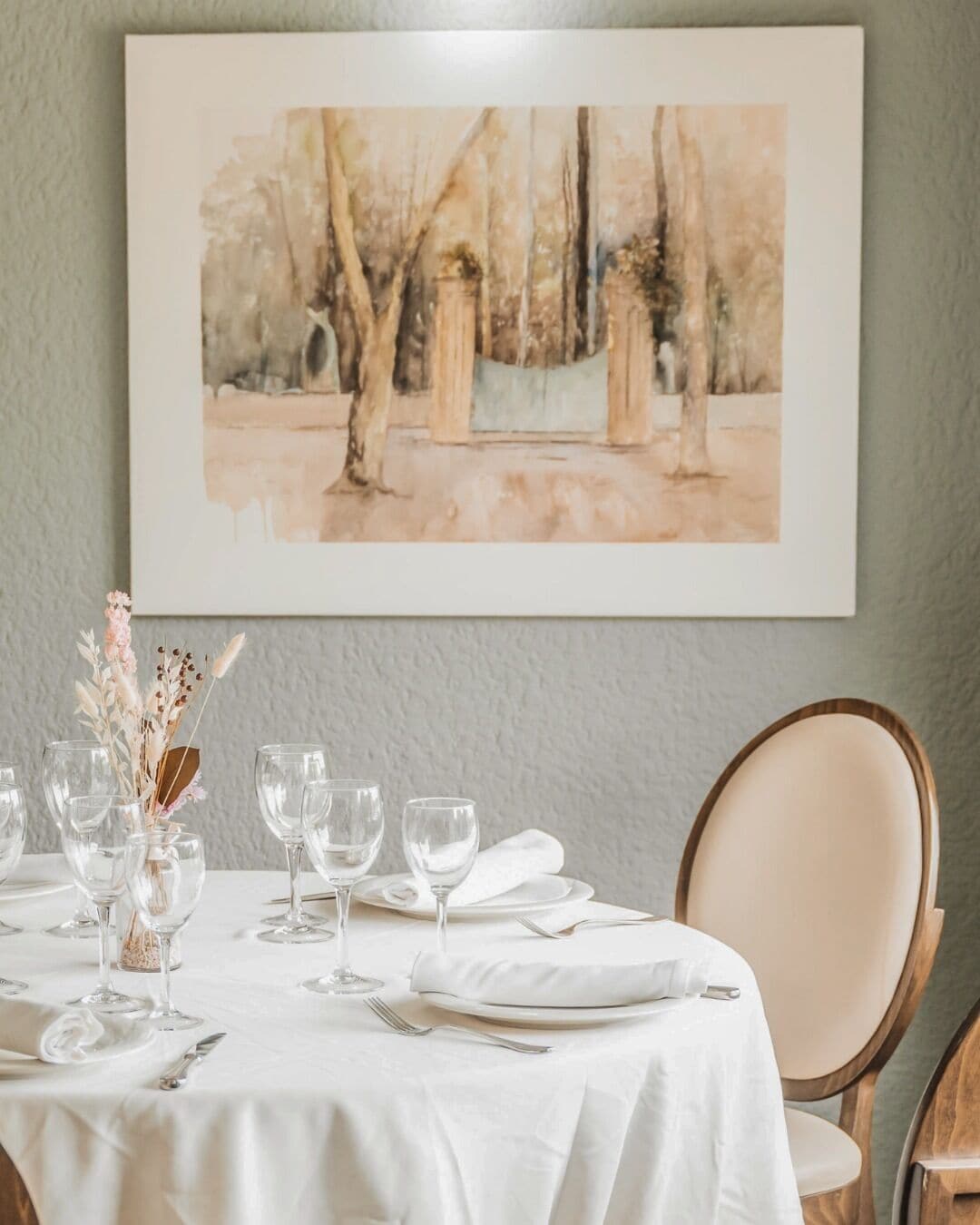 Una mesa de comedor dispuesta con mantel blanco, copas de vino y un florero con flores secas.