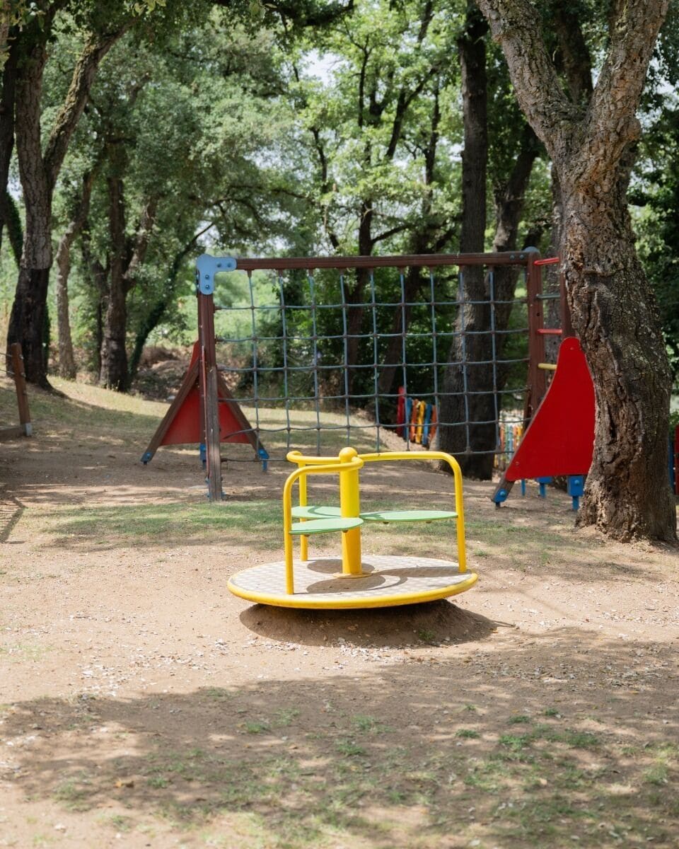 A playground with a yellow merry-go-round, a red climbing structure, and a netted play area surrounded by trees.