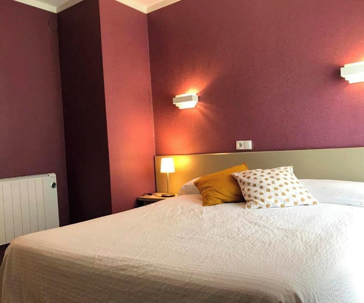 Et hotelværelse med seng, natbord, lampe og vægmonteret lys.