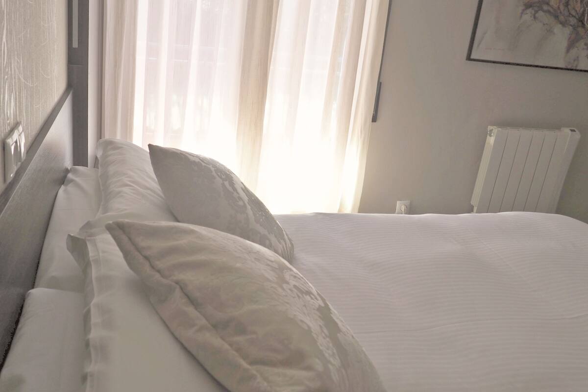 Uma cama bem arrumada com lençóis e almofadas brancas, uma janela com cortinas leves e um radiador na parede.