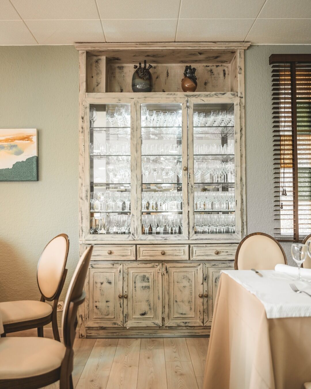 Un comedor con un armario de madera que exhibe cristalería, una mesa dispuesta para dos personas y una ventana con persianas.