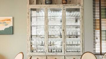 Un comedor con un armario de madera que exhibe cristalería, una mesa dispuesta para dos personas y una ventana con persianas.