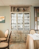 Un comedor con un armario de madera que exhibe cristalería, una mesa dispuesta para dos personas y una ventana con persianas.