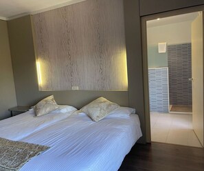A modern hotel room with a large bed, two pillows, a nightstand, and a wall-mounted light fixture.