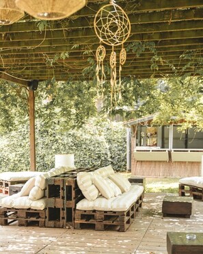 A covered outdoor seating area with a wooden bench and cushions, surrounded by greenery and a bamboo fence. - Hostal Esteba (Caldes de Malavella)