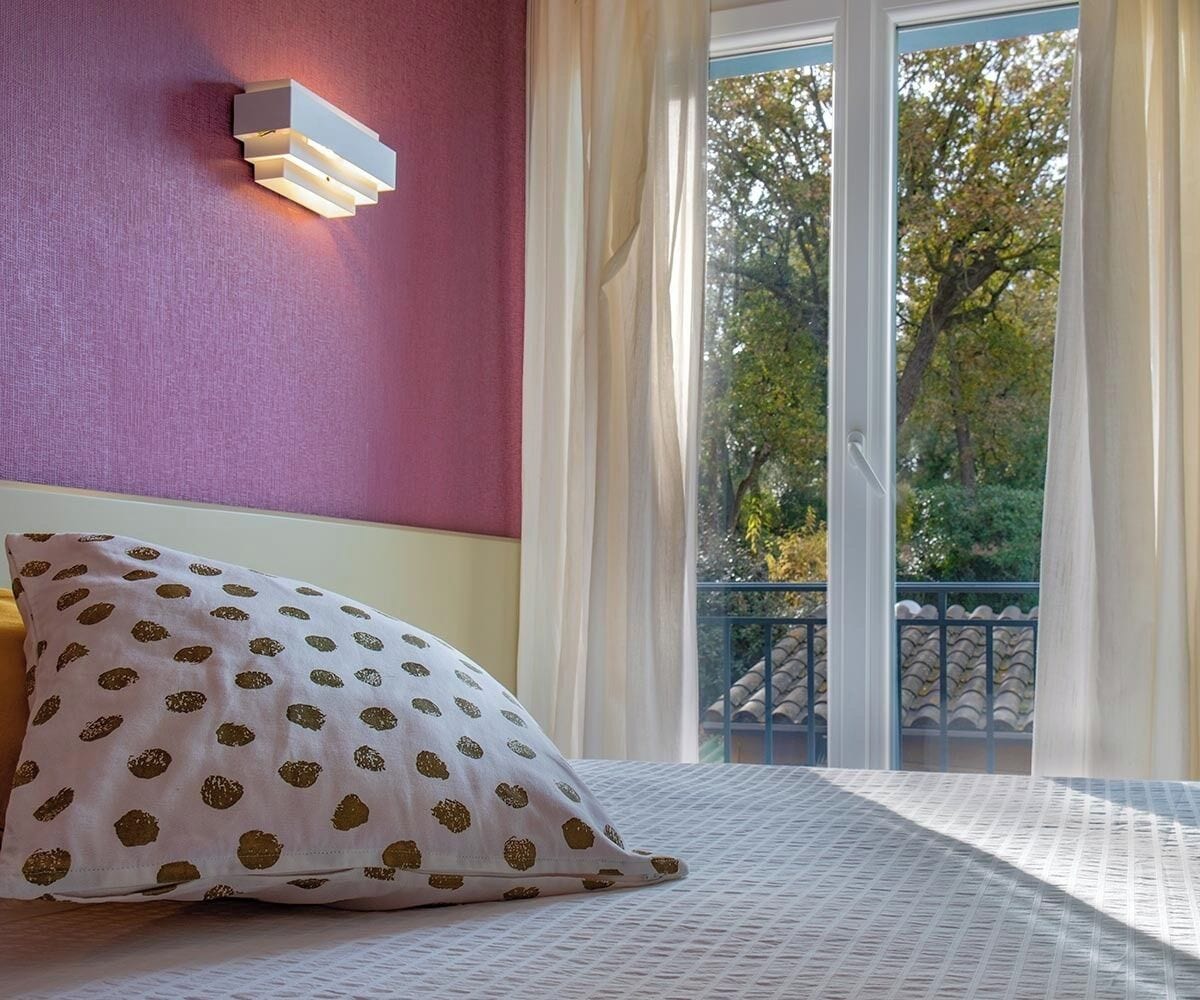 Un dormitorio con cama, lámpara de pared, ventana con cortinas y vista a árboles.