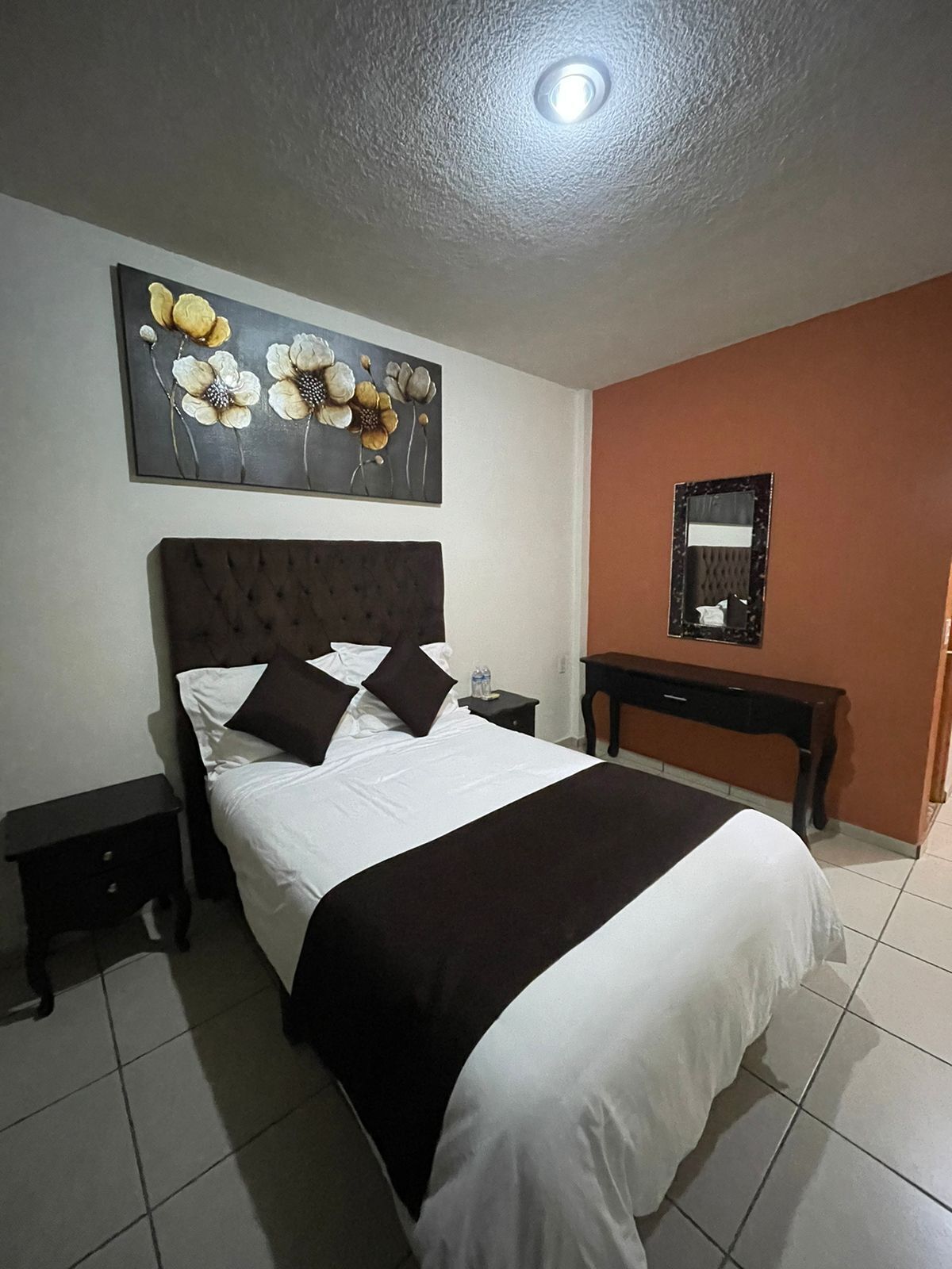 Cama bem arrumada com cabeceira escura, mesa de cabeceira, espelho fixado na parede e uma obra de arte floral acima da cama.