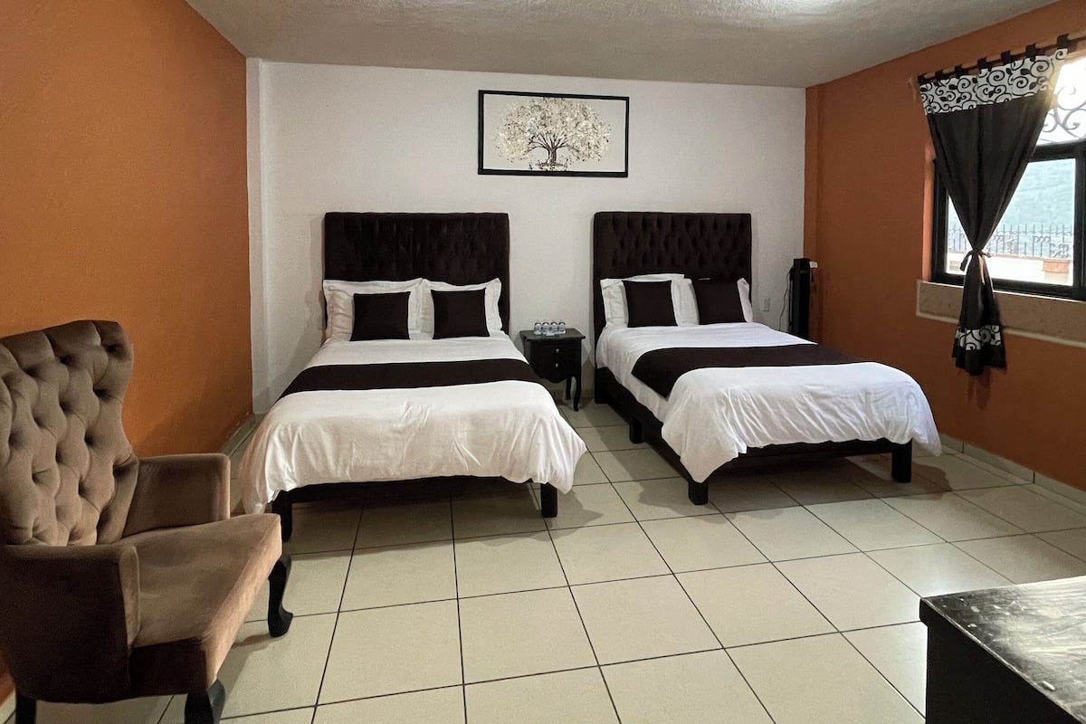 A hotel room with two beds, a chair, a framed picture on the wall, and a window with curtains.