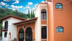 Un edificio de dos plantas con fachada color beige y techo de tejas rojas. Se ven dos ventanas en la planta baja y un balcón con barandilla.