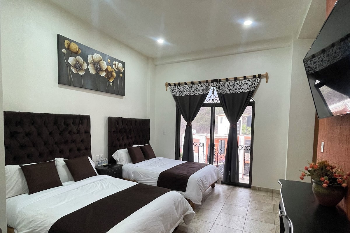 A hotel room with two beds, a flat-screen TV, a painting, and a dining table with a flower pot.