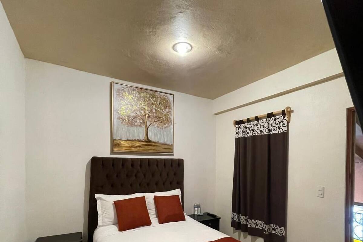 A bedroom with a large bed, a headboard, a nightstand, a painting on the wall, and a window with curtains.