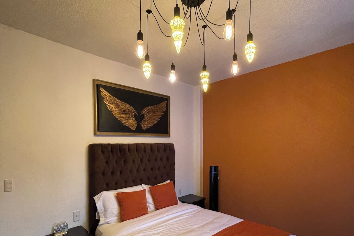 A bedroom with a tufted headboard, a large bed with white and orange bedding, a wall-mounted light fixture, and a framed artwork above the bed.