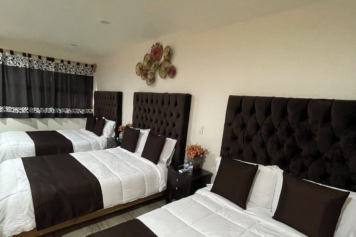 A hotel room with three beds, each with a tufted headboard, white bedding, and dark brown accents. There is a decorative wall piece above the beds and a small table with a vase of flowers.