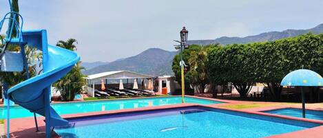 A swimming pool with a blue slide, a covered seating area, and a mountain view in the background.