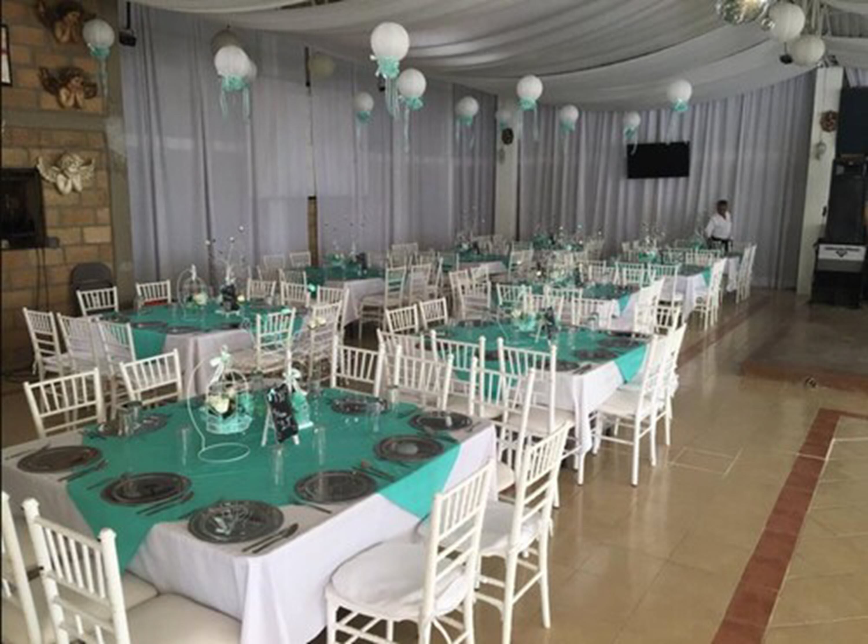A banquet hall with multiple round tables covered in white tablecloths and teal table runners, surrounded by white chairs.