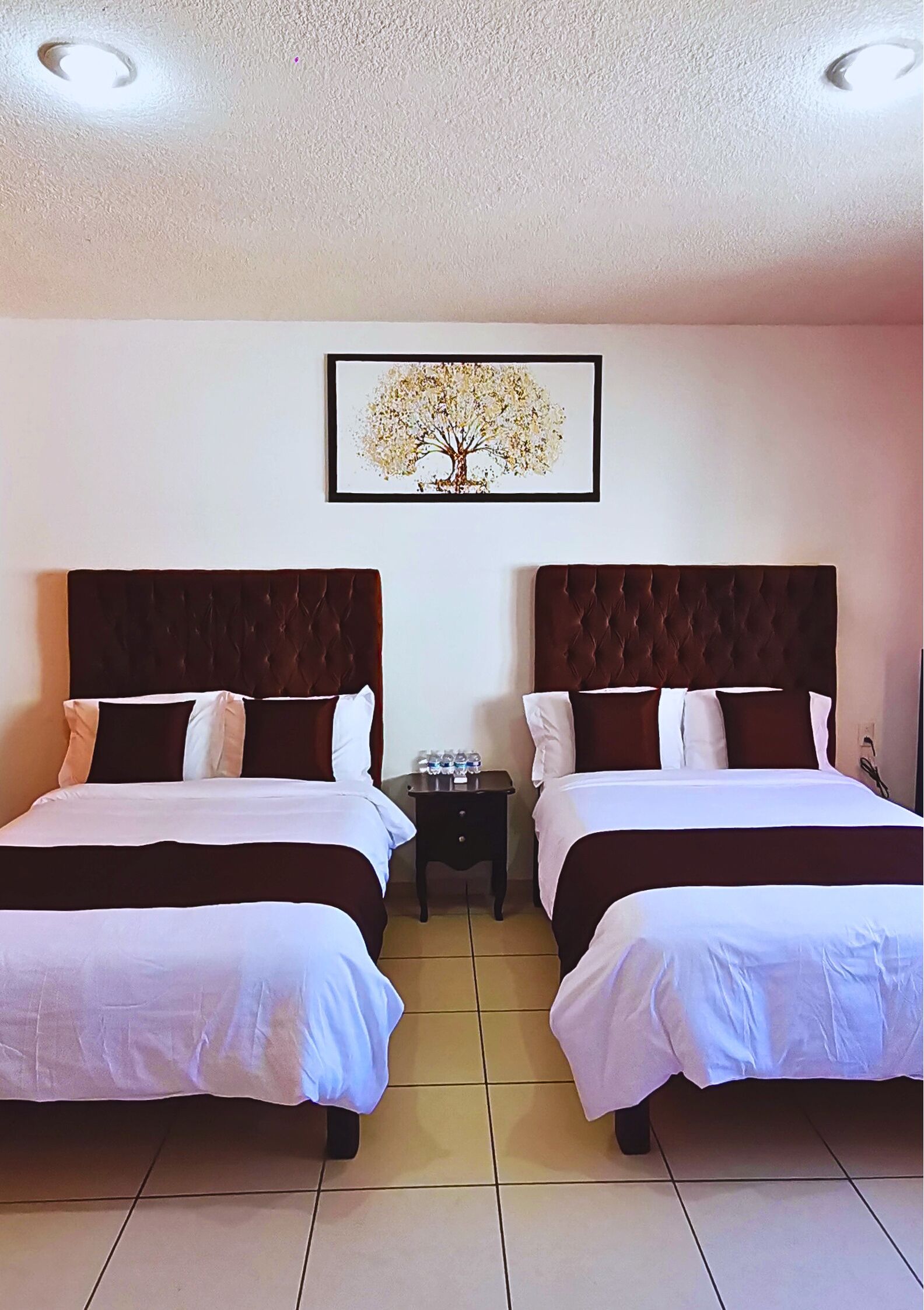 A hotel room with two beds, each with a dark headboard and white bedding. There is a framed picture of a tree on the wall above the beds.