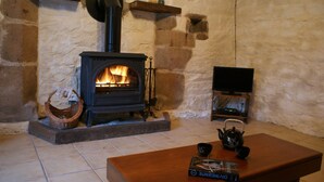 A stone-walled room with a wood-burning stove, a television on a stand, and a table with a book and a teapot. - 3 houses with 6x3 beds, approx. 1 hectare of land and a swimming pool. (Ploërdut)