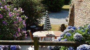 A patio area with a round wooden table and chairs, surrounded by flowering plants and a view of a body of water. - 3 houses with 6x3 beds, approx. 1 hectare of land and a swimming pool. (Ploërdut)