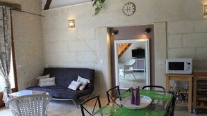 A cozy room with a dark blue sofa, a glass-top dining table with chairs, a white wicker chair, a clock on the wall, and a microwave on a wooden shelf. - Lodging near Amboise, Chenonceau, Chaumont / Loire  (Mosnes)