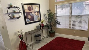 Un salon avec un tapis rouge, une plante en pot, une console, un tableau abstrait encadré et une étagère murale agrémentée d’objets décoratifs.