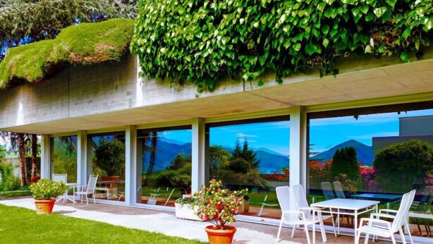 Un bâtiment moderne doté d’une façade en verre, d’un espace extérieur aménagé pour s’asseoir et offrant une vue sur les montagnes.