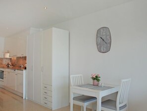 Moderni keittiö, jossa on valkoiset kaapit, ruokailutila valkoisella pöydällä ja tuoleilla sekä seinälle kiinnitetty kello.