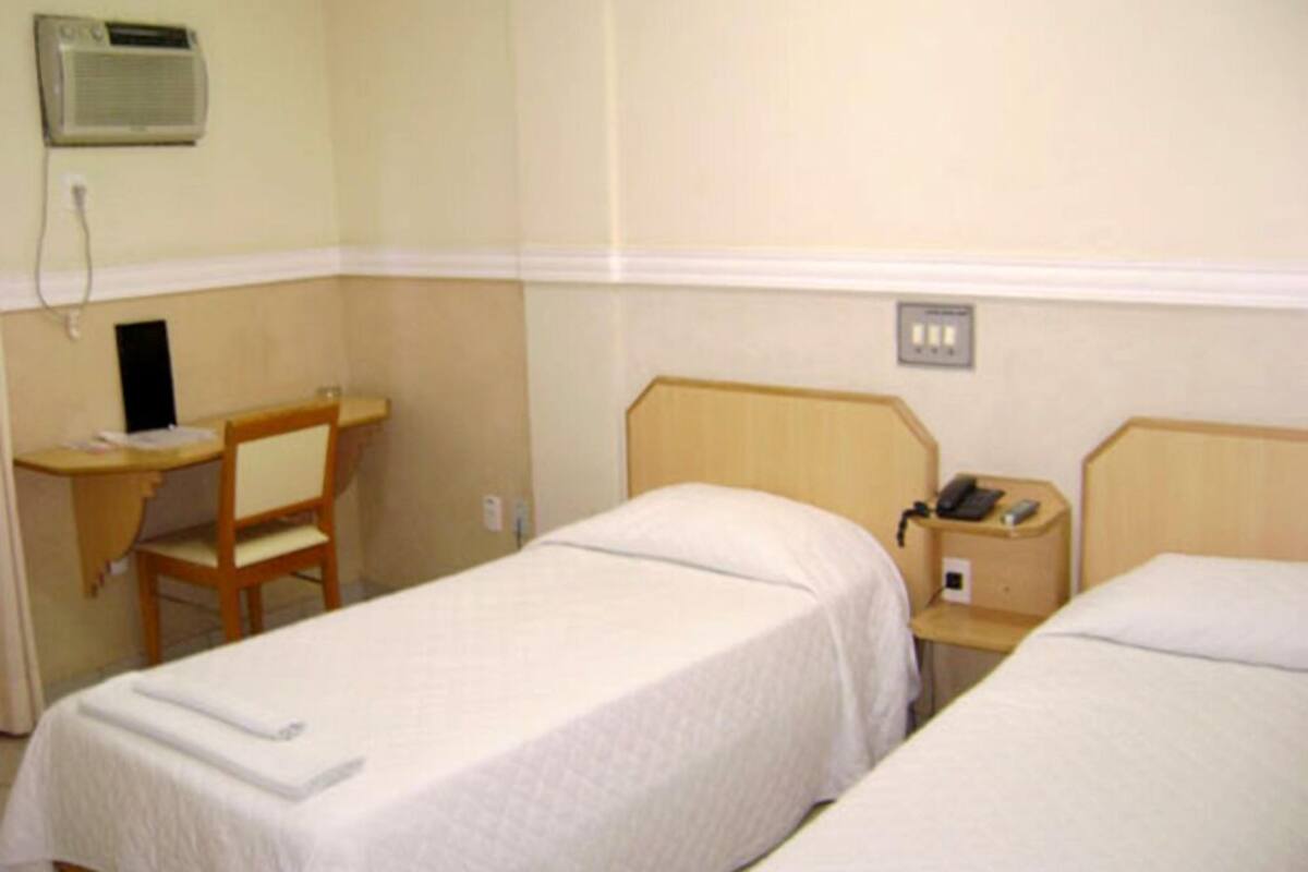 Une chambre d’hôtel avec deux lits simples, un bureau avec une chaise, une table de chevet avec un téléphone et un appareil de climatisation fixé au mur.