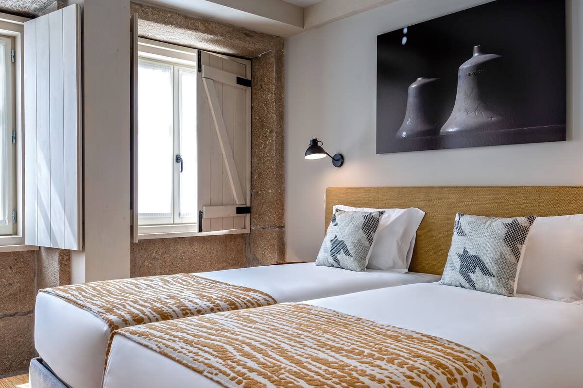 A hotel room with two beds, a large painting, a wall-mounted lamp, and a window with curtains.