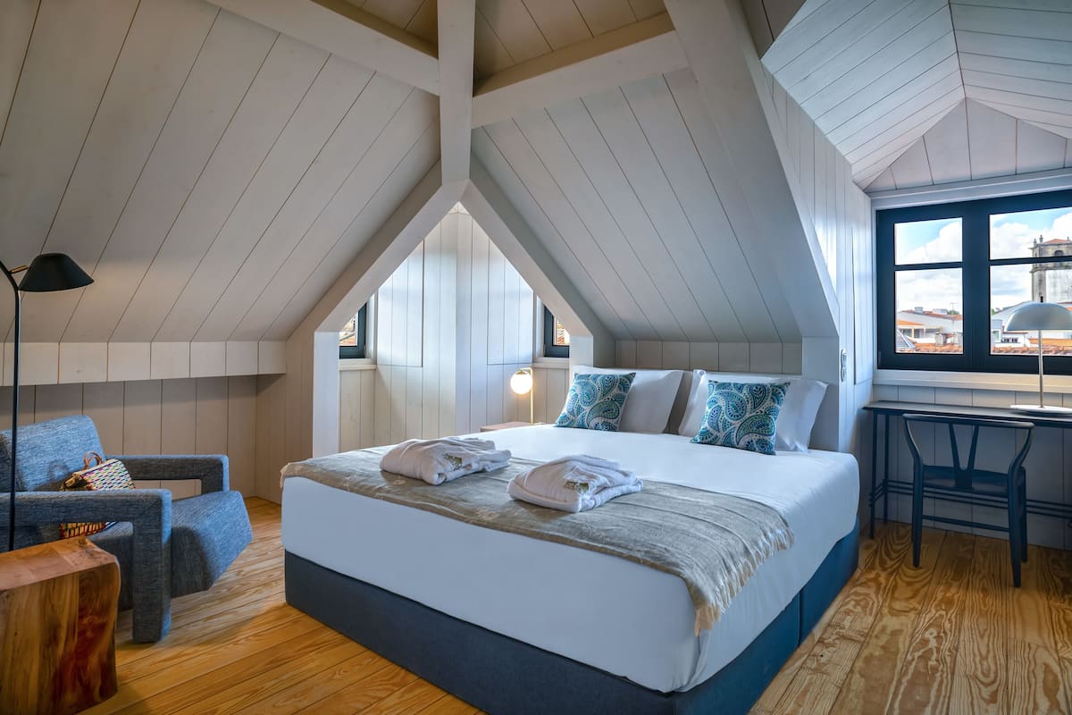 A bedroom with a high ceiling, a bed with white linens, a blue chair, and a wooden floor.