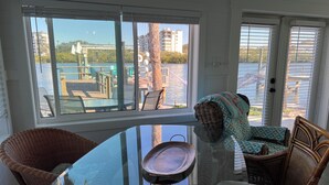 A room with a glass table and wicker chairs, a view of a waterfront with buildings and greenery.