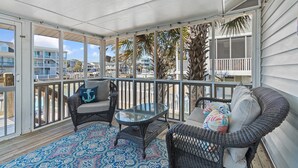 Een balkon met overkapping, met rieten meubels, een glazen tafel en een kamerkleed met patroon.