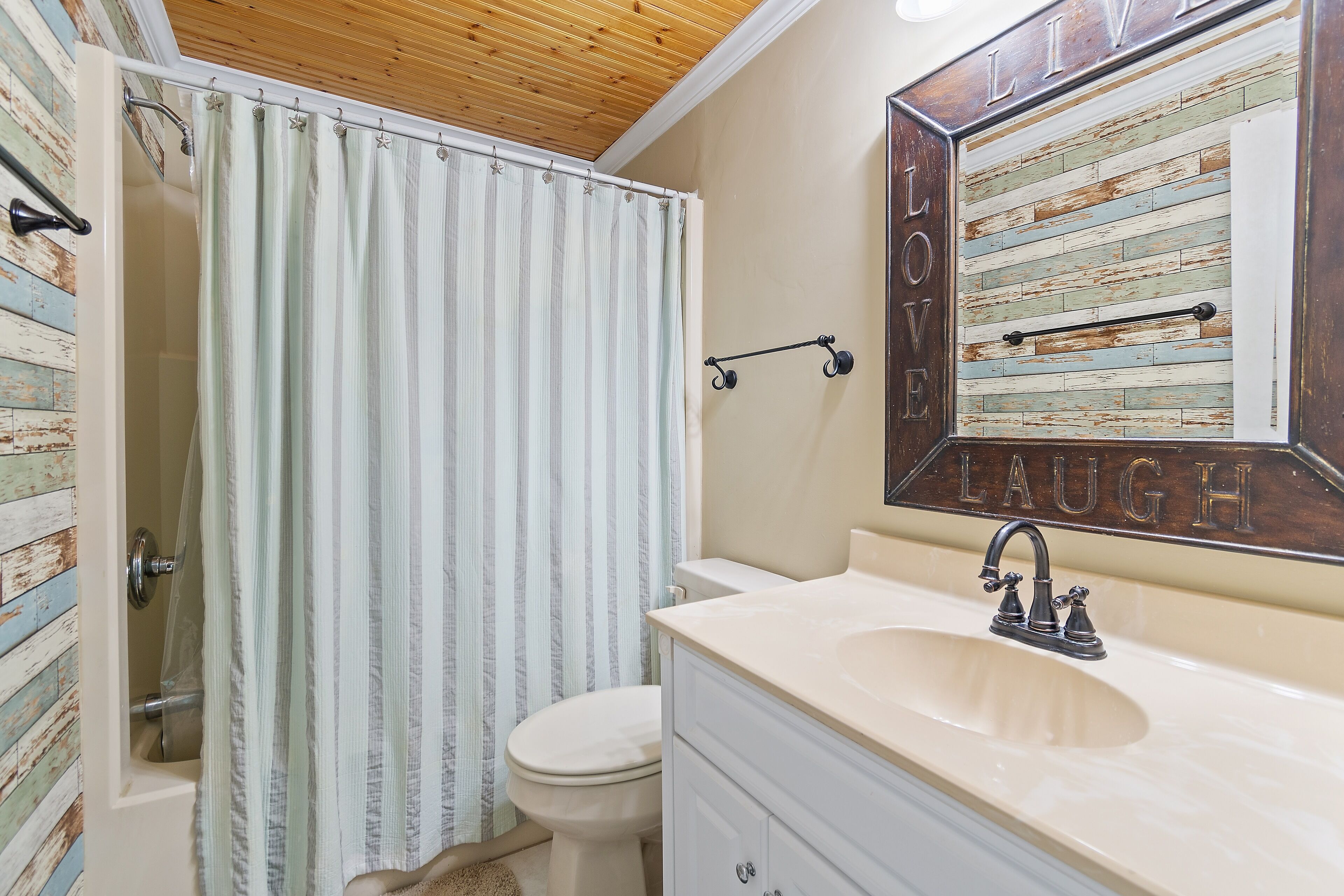 Une salle de bain avec un plafond en bois, un rideau de douche à rayures, des toilettes, un lavabo avec un miroir et un mur sur lequel on peut lire les mots « LOVE LAUGH ».