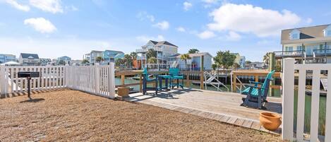 A wooden deck with outdoor seating, a white fence, and a view of the ocean and buildings in the background.