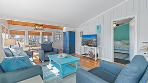 Een woonkamer met een blauwe bank, een bijpassende salontafel en een televisie. Er is een houten plafond en uitzicht op het buitenland door grote ramen.