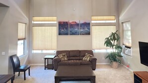 A spacious living room with a brown sofa, a coffee table, and a large window with blinds. There are two wall-mounted paintings and a potted plant. - Mountain Retreat - Close to NAU & Snowbowl (Bellemont)