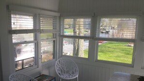 A room with a view of a green lawn and a tree, a white chair, a small table, and a window with blinds. - Adorable Cottage Townline Lake Paradise. Boat Kayak Fish BBQ Relax Swim. 2 BR  (Lakeview)