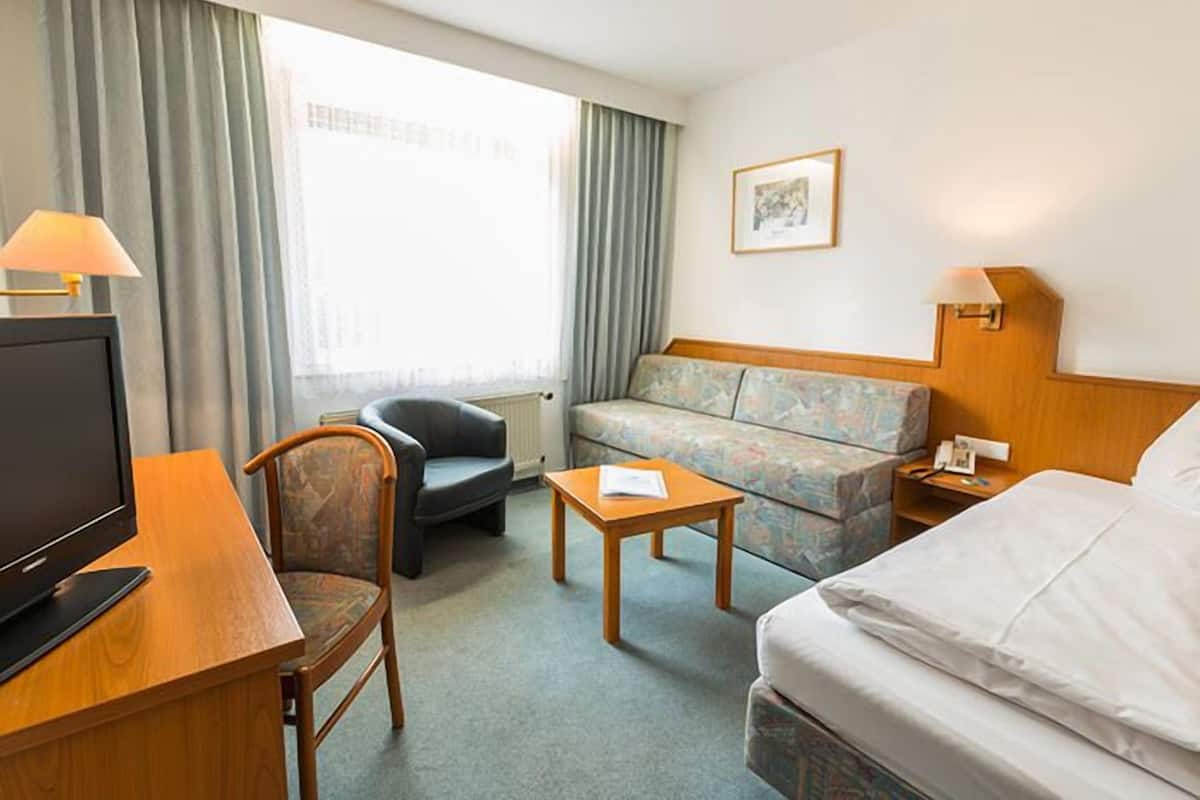 A hotel room with a bed, a sofa, a desk with a computer, a chair, a small table, and a window with curtains.