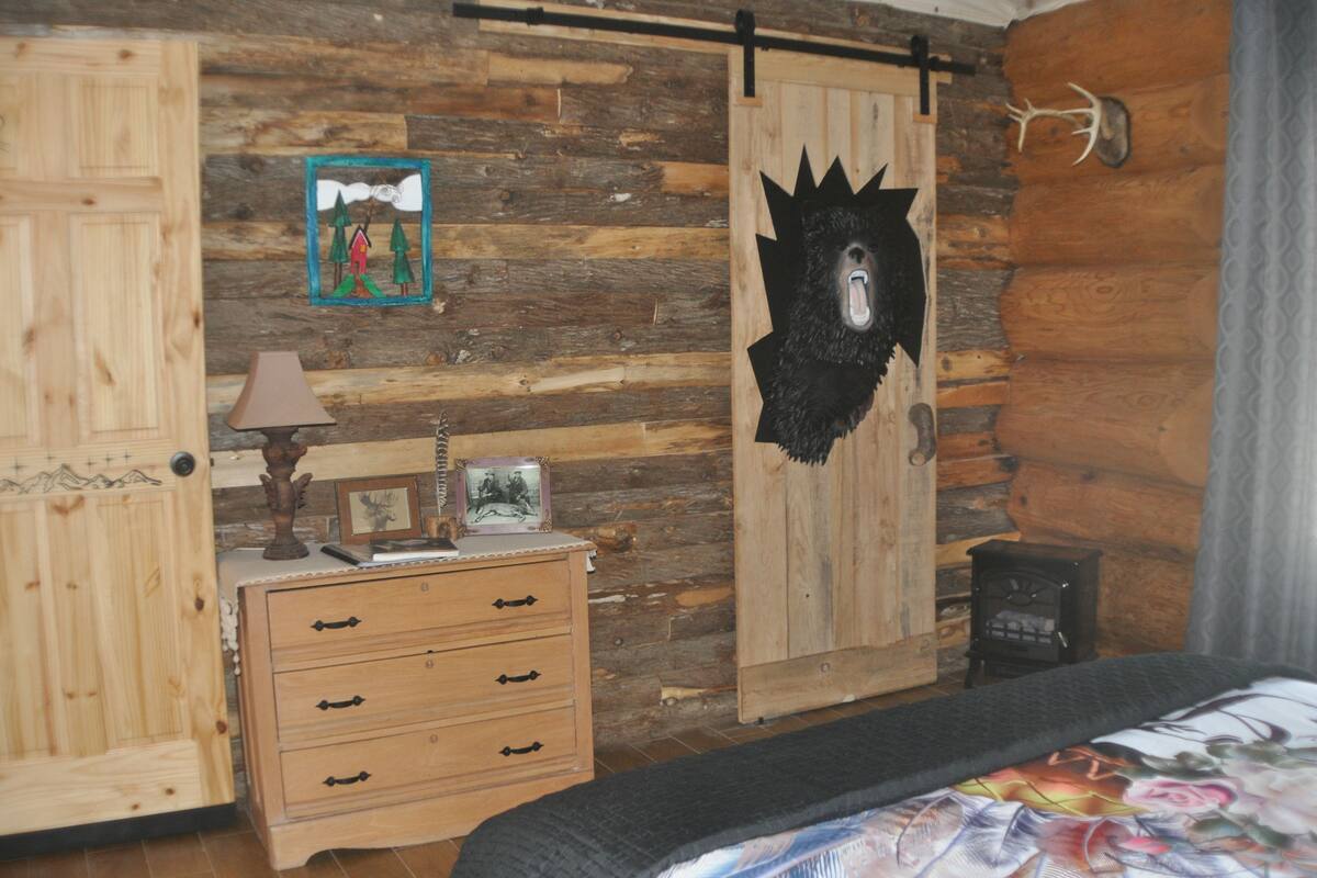 A rustic cabin room with wooden walls, a bed with a floral bedspread, a dresser with drawers, a lamp, and a framed picture.