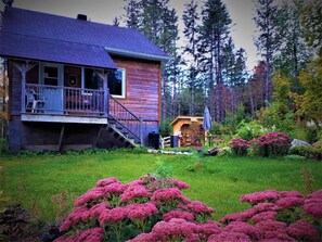 Una cabaña de madera con porche y una escalera que conduce a una terraza, rodeada de un exuberante jardín y altos árboles.