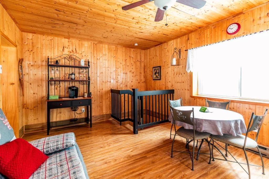 A wooden-paneled room with a dining table, chairs, a sofa, a small table with a coffee machine, and a ceiling fan.