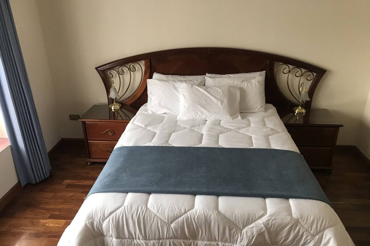 Ein ordentlich bezogenes Bett mit weißer Steppdecke und blauer Decke, begleitet von zwei Nachttischen mit Lampen und einem Spiegel.